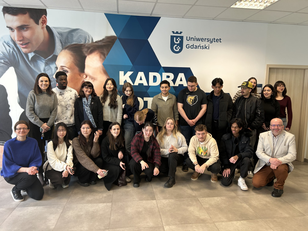 A group of Erasmus+ students pose for a group photo against the background of a wall with the University of Gdańsk logo
